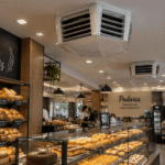 Interior de uma padaria moderna com prateleiras de pães, balcão de vidro com doces e salgados, atendente e clientes sentados às mesas ao fundo.